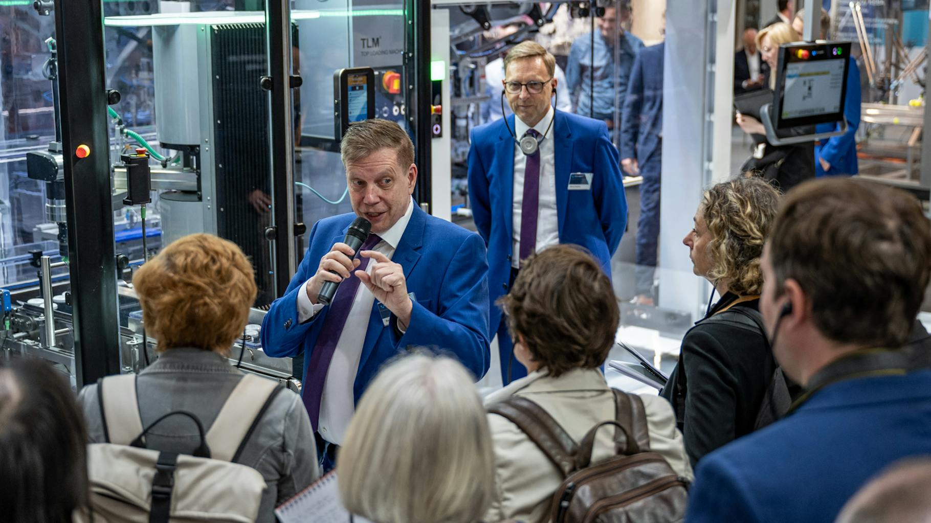 A crowd listens to a speaker from pr agency schubert talking into a microphone and standing in front of various machines.