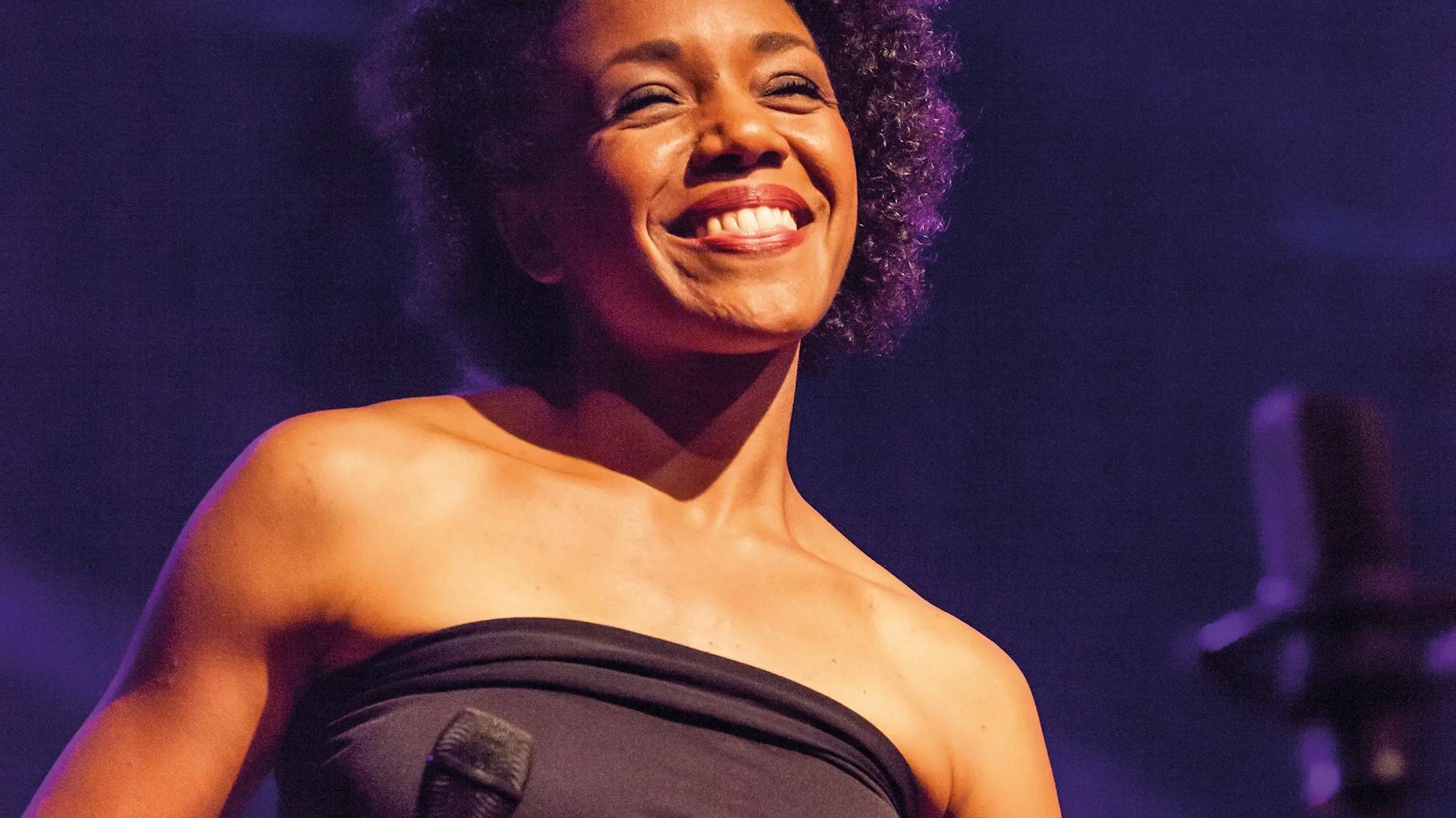 A backing singer, illuminated by the stage lights, smiles at the crowd.