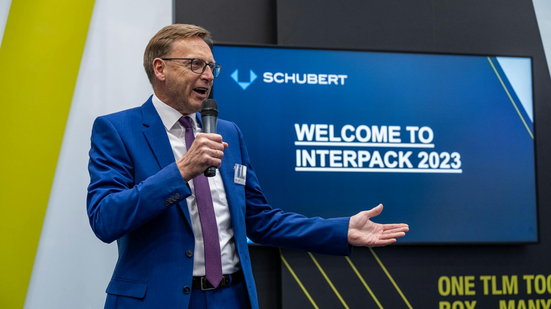 Ein Sprecher der pr agentur schubert spricht in ein Mikrofon und steht vor einem Monitor, auf dem geschrieben steht "welcome to interpack 2023".