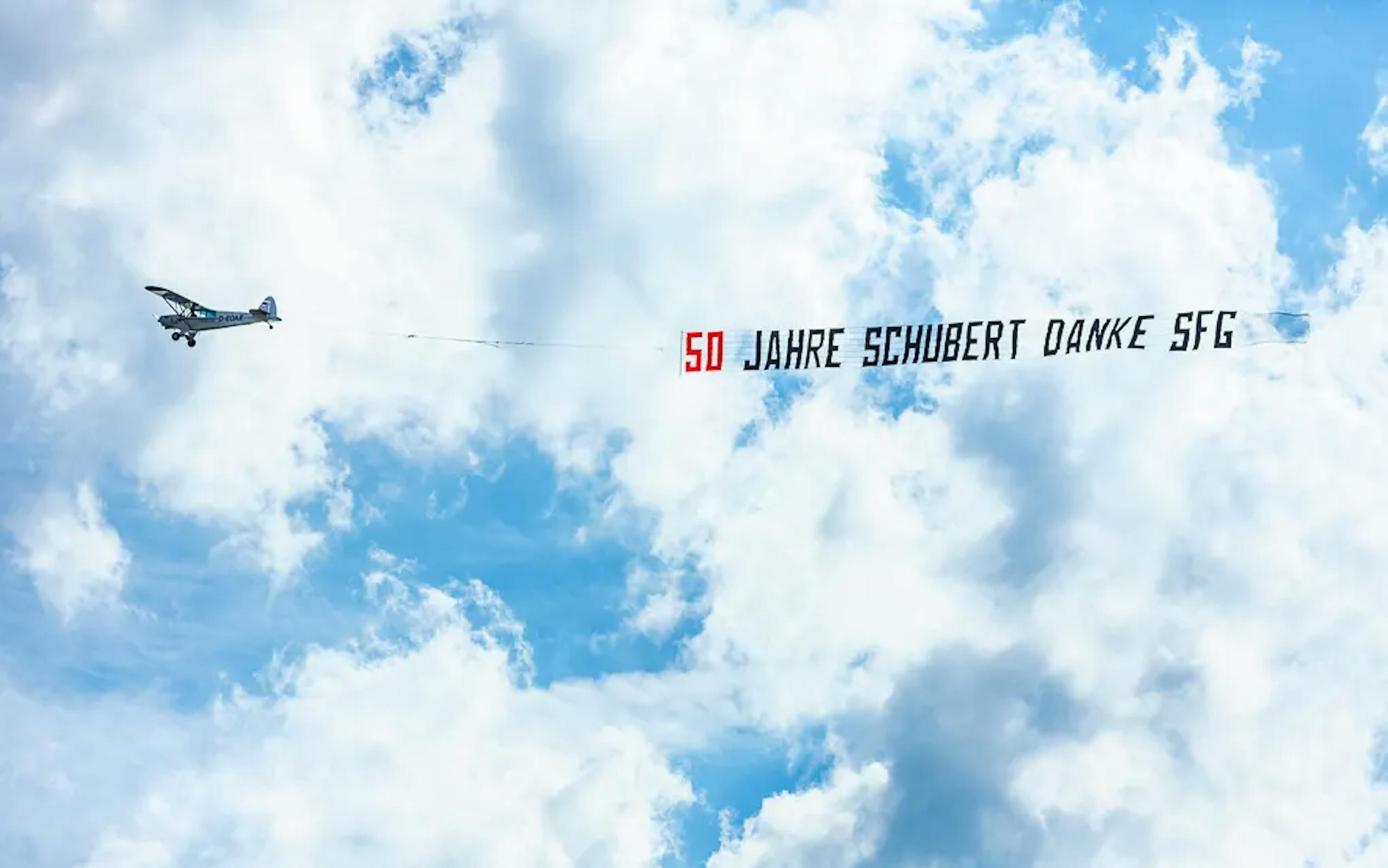 Ein Flugzeug ist vor Wolken zu sehen, es zieht einen Banner mit der Aufschrift "50 Jahre Schubert Danke SFG" hinter sich her.