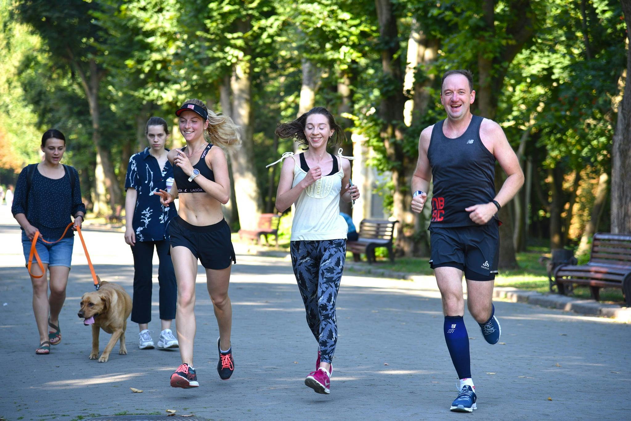 На зображенні - три учасники забігу Runday усміхнені біжать через парк, оминаючи пішоходів