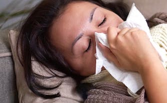 person with covid-19 lying on couch, wrapped in blanket, and blowing nose