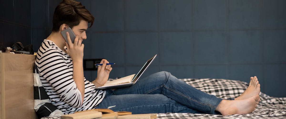teenager talking on phone with computer and notebook open in bed
