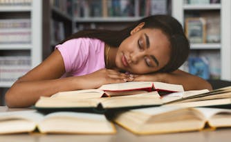 college student sleeping on top of pile of books
