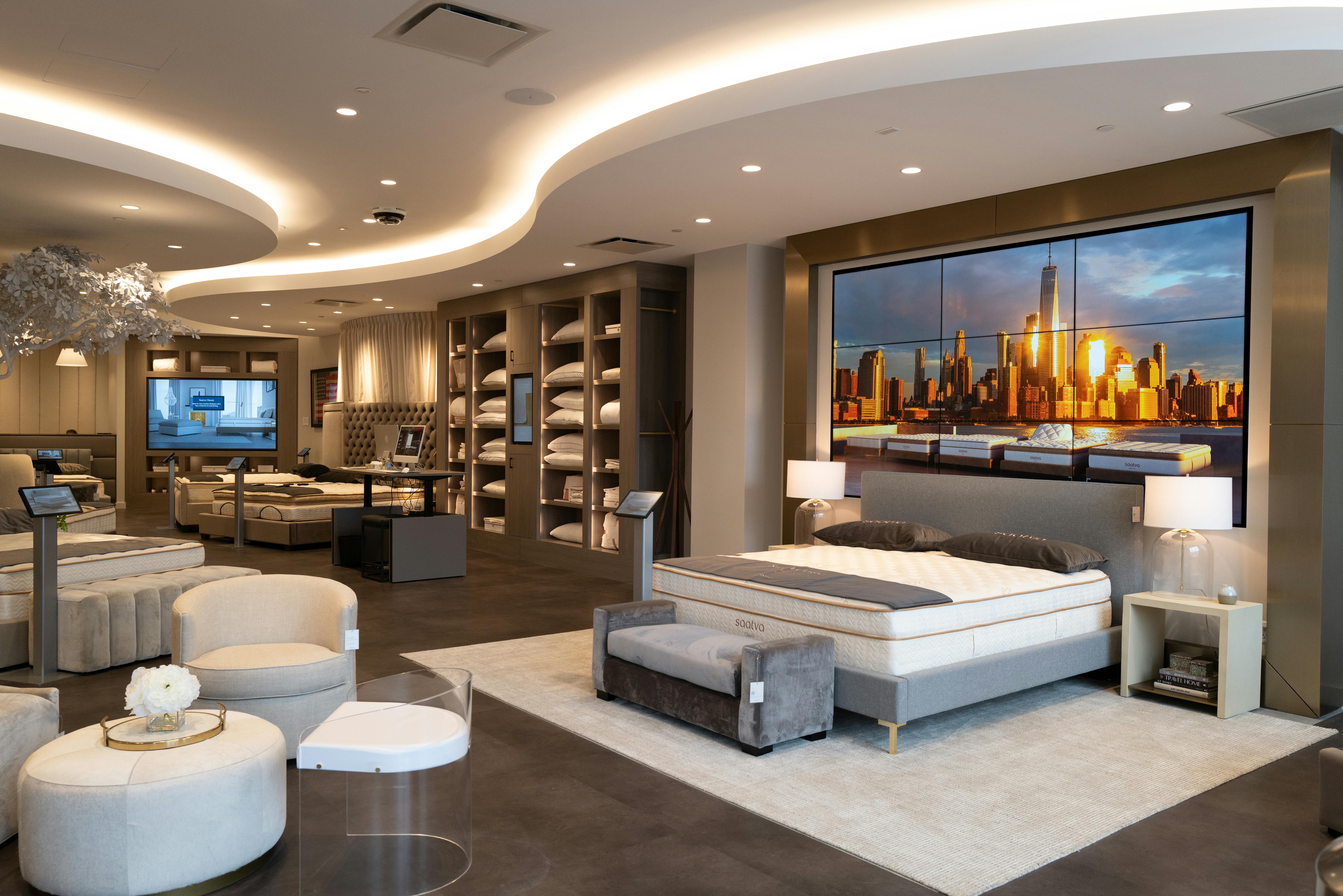 The elegant interior of the Saatva Manhasset retail location features a bed with a digital headboard, a self-checkout station with desktop computers for guests to transact at their leisure, a bedding and sleep accessories wall, and a seating area.