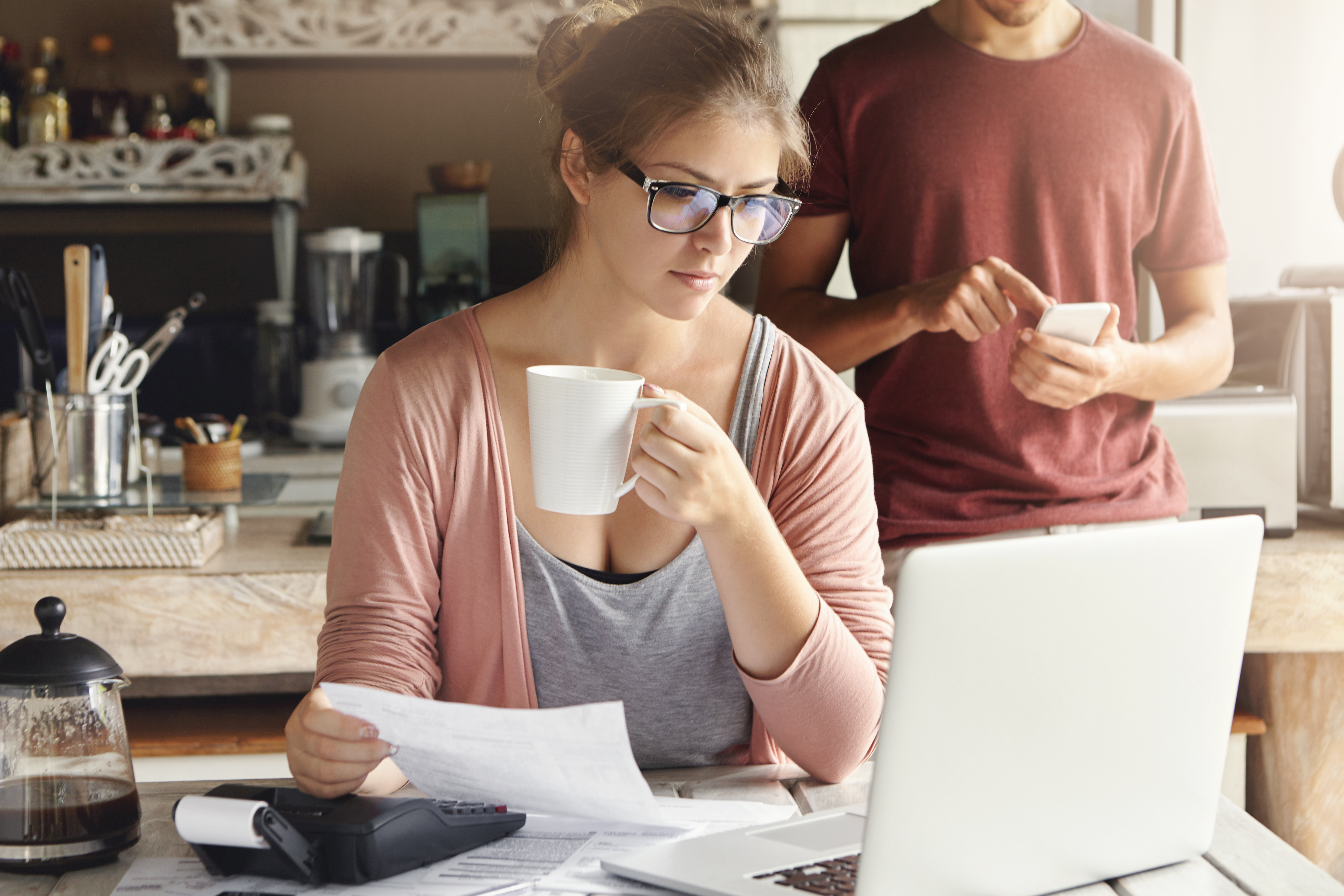 Young female having concentrated expression looking at screen of open laptop, holding paper and cup of coffee in her hands
