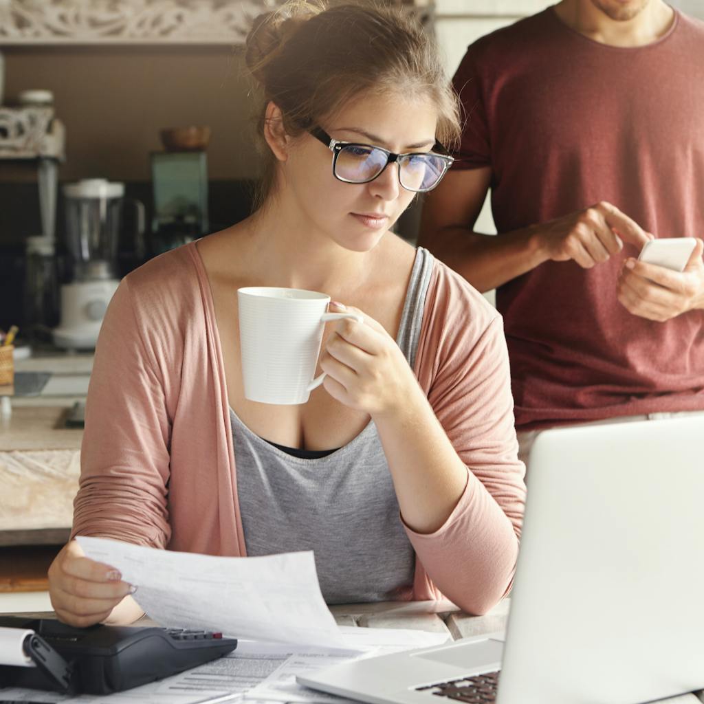 Young female having concentrated expression looking at screen of open laptop, holding paper and cup of coffee in her hands