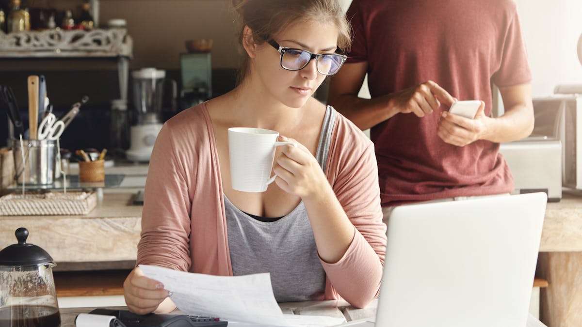 Young female having concentrated expression looking at screen of open laptop, holding paper and cup of coffee in her hands