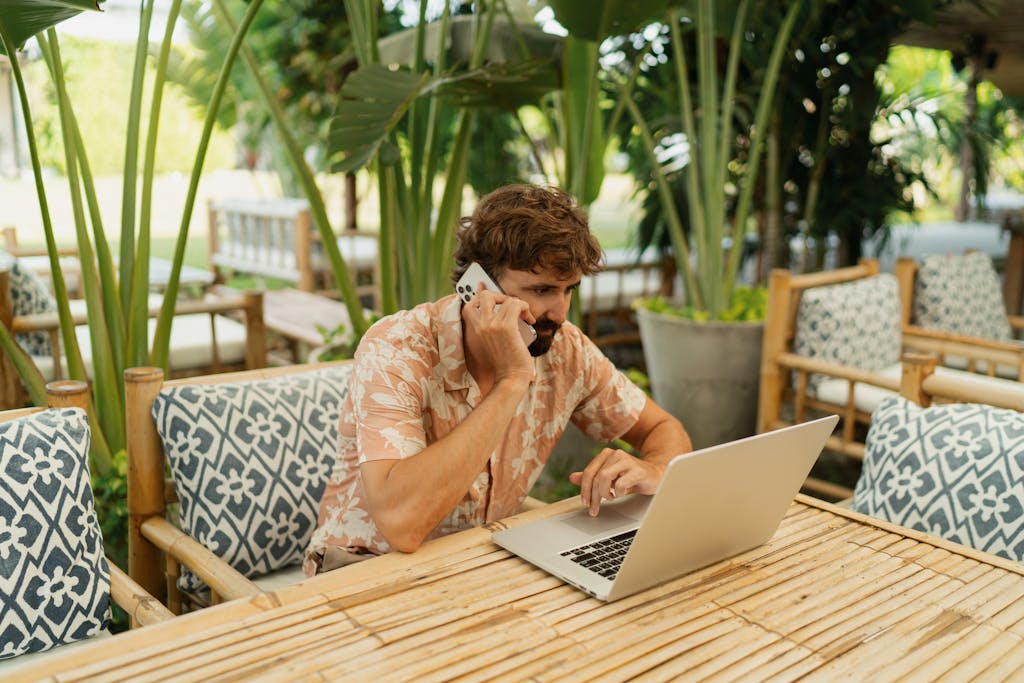 Man with beard using lap top and mobile phone sitting in outdoor cafe with tropical interior