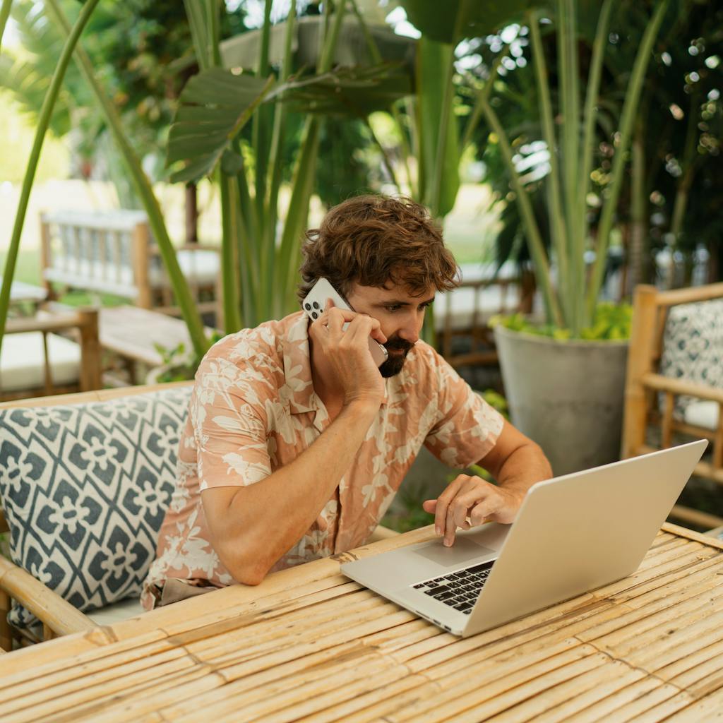 Man with beard using lap top and mobile phone sitting in outdoor cafe with tropical interior