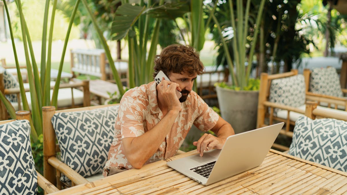 Man with beard using lap top and mobile phone sitting in outdoor cafe with tropical interior