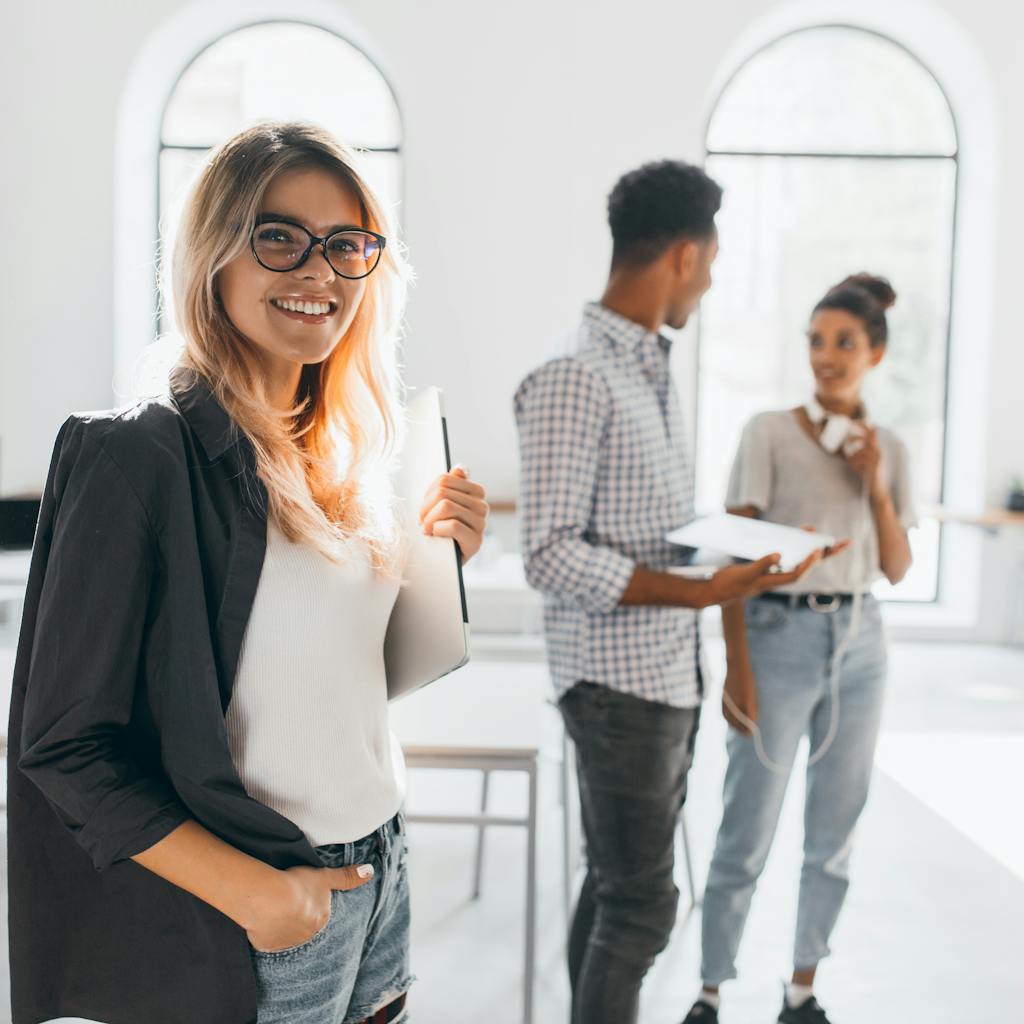 Dama elegante de negocios con chaqueta negra a la moda sosteniendo un portátil y sonriendo en una oficina de aspecto moderno.