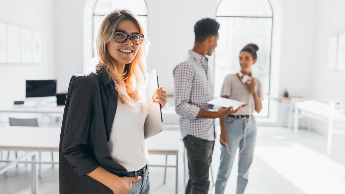 Dama elegante de negocios con chaqueta negra a la moda sosteniendo un portátil y sonriendo en una oficina de aspecto moderno.