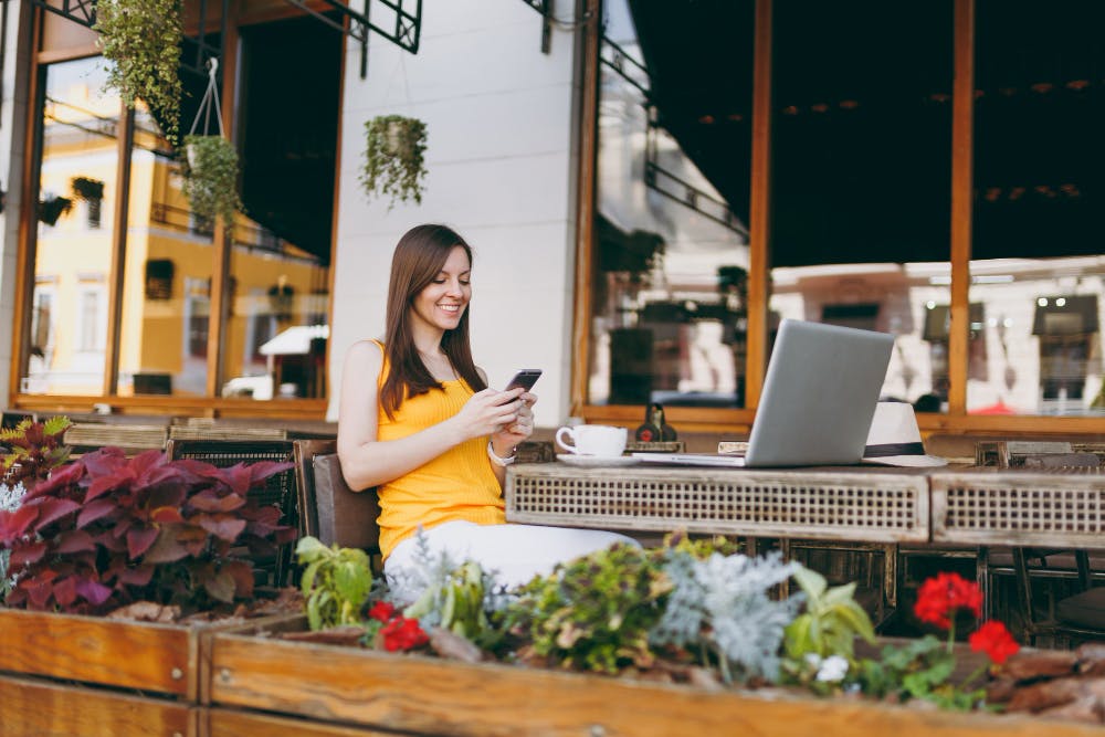 Happy smiling girl in outdoors street coffee shop cafe sitting at table with laptop pc computer, texting message on mobile phone friend, in restaurant during free time <a href="https://www.freepik.com/photos/business">Business photo created by ViDIstudio - www.freepik.com</a>