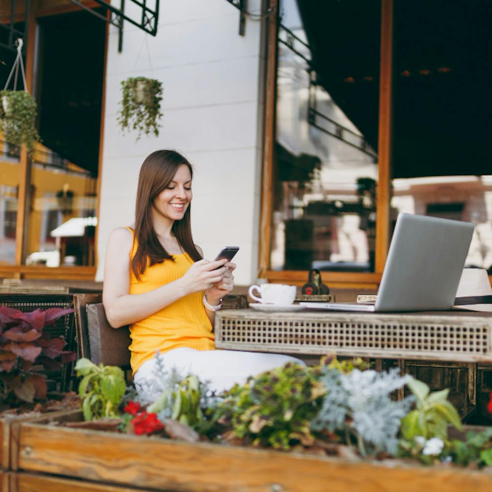 Happy smiling girl in outdoors street coffee shop cafe sitting at table with laptop pc computer, texting message on mobile phone friend, in restaurant during free time <a href="https://www.freepik.com/photos/business">Business photo created by ViDIstudio - www.freepik.com</a>