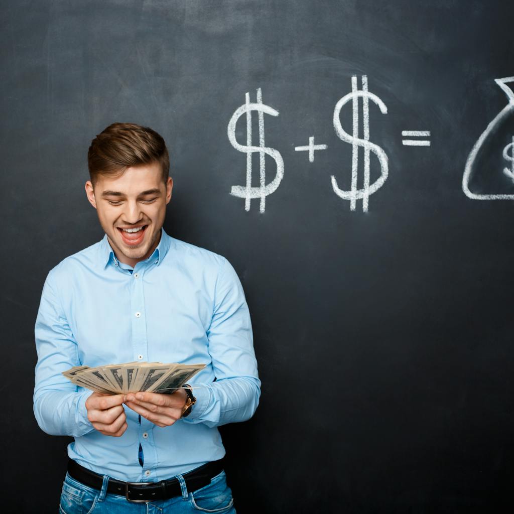 Man holding several bills of U.S. dollars standing over a blackboard with drawn dollar signs 