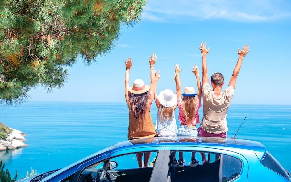 A family at the top of car raising their hands in front of the sea