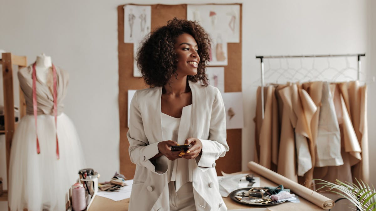 Curly-haired brunette woman in oversized jacket and white pants smiles, holds phone and leans on table in office