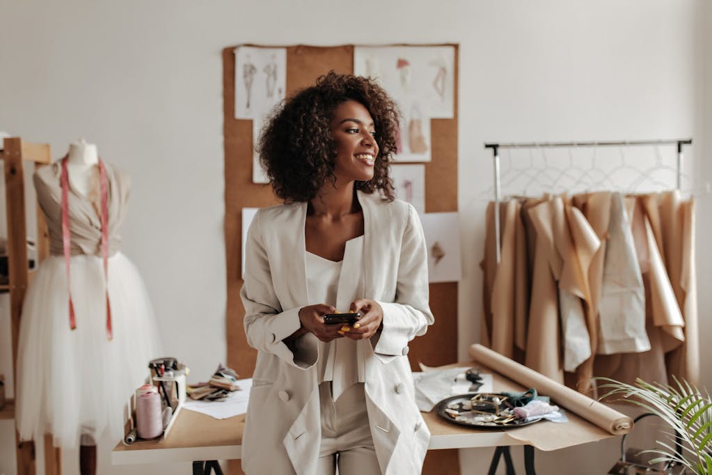Curly-haired brunette woman in oversized jacket and white pants smiles, holds phone and leans on table in office