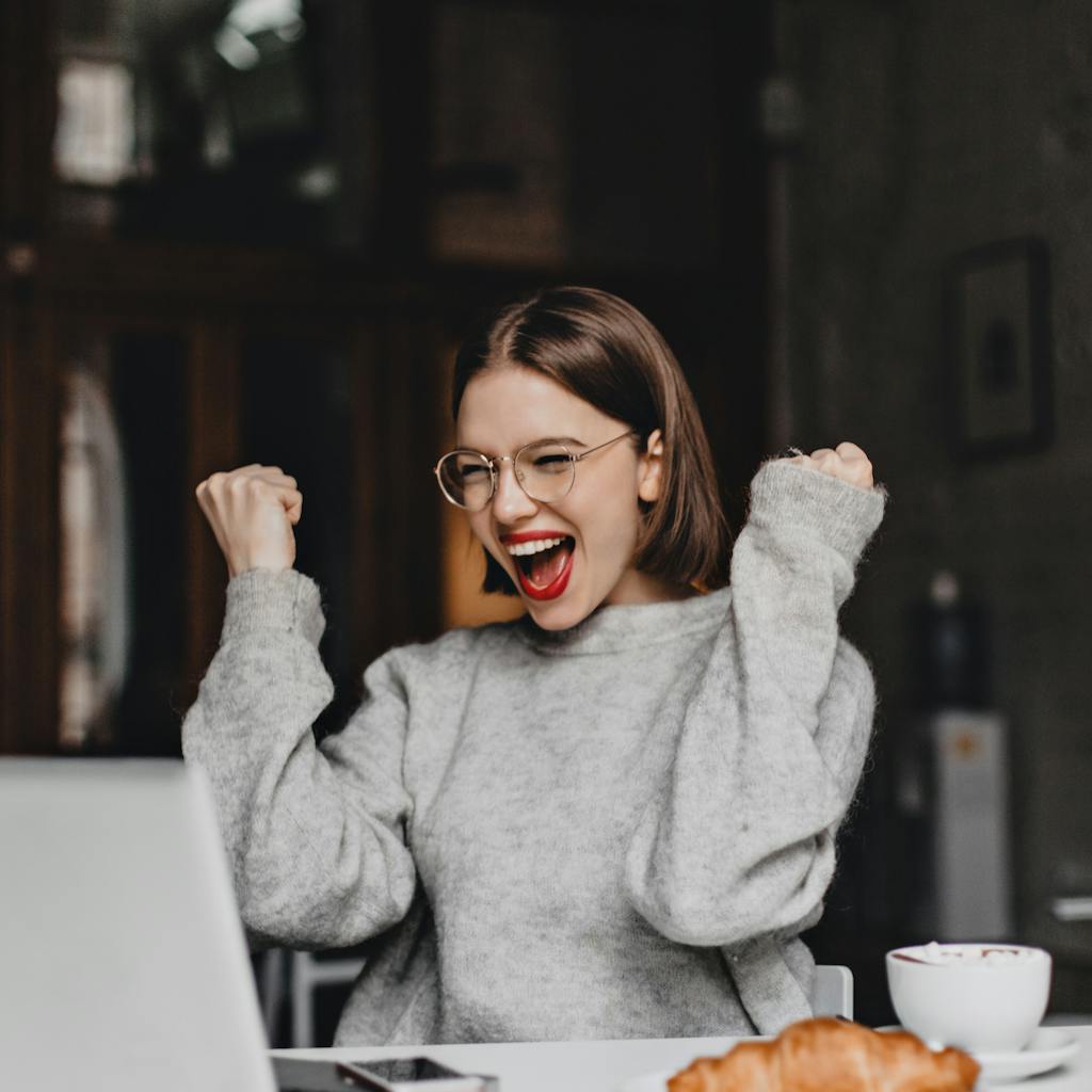 Happy woman in glasses makes winning gesture and sincerely rejoices. lady with red lipstick dressed in gray sweater looking at laptop. <a href="https://www.freepik.com/photos/coffee">Coffee photo created by lookstudio - www.freepik.com</a>