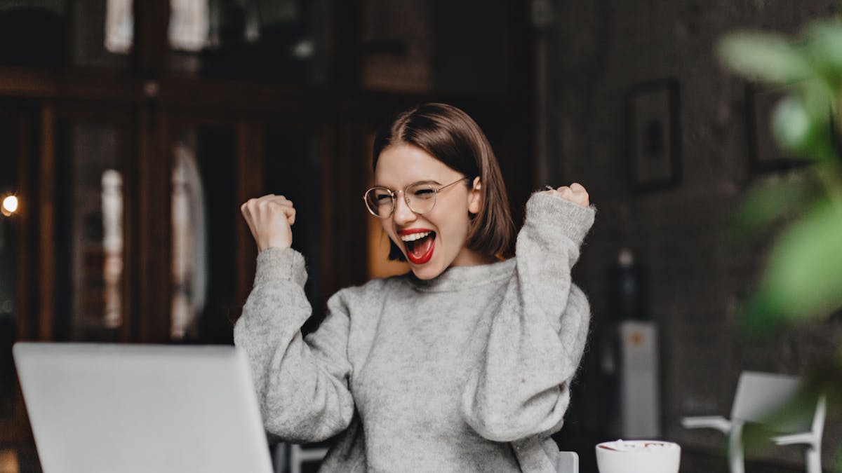 Happy woman in glasses makes winning gesture and sincerely rejoices. lady with red lipstick dressed in gray sweater looking at laptop. <a href="https://www.freepik.com/photos/coffee">Coffee photo created by lookstudio - www.freepik.com</a>