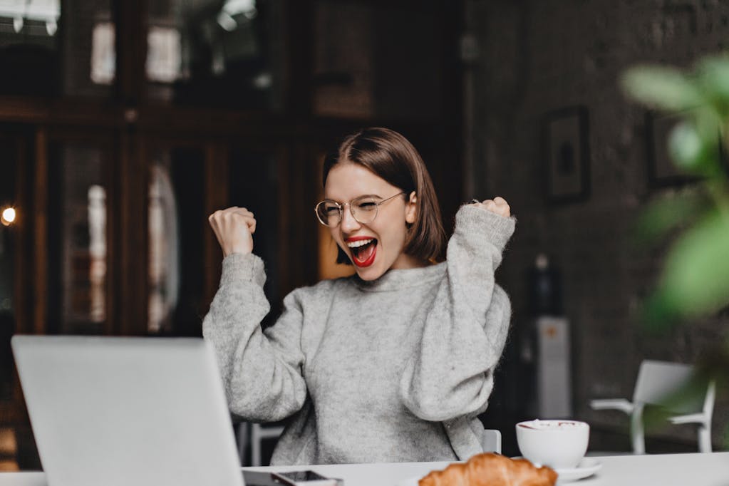 Happy woman in glasses makes winning gesture and sincerely rejoices. lady with red lipstick dressed in gray sweater looking at laptop. <a href="https://www.freepik.com/photos/coffee">Coffee photo created by lookstudio - www.freepik.com</a>