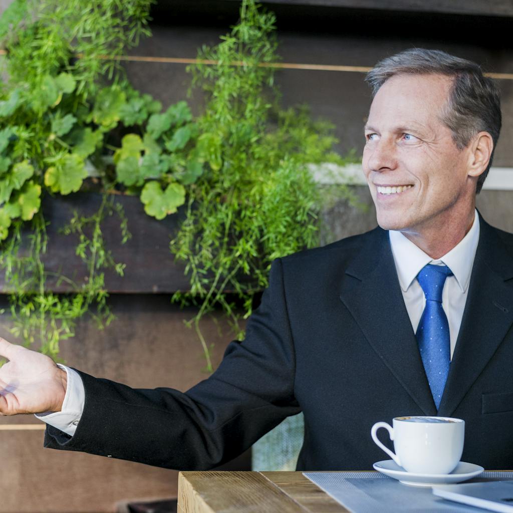 Smiling mature businessman making hand gesture with cup of coffee and laptop on desk