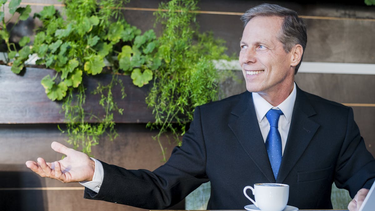 Smiling mature businessman making hand gesture with cup of coffee and laptop on desk