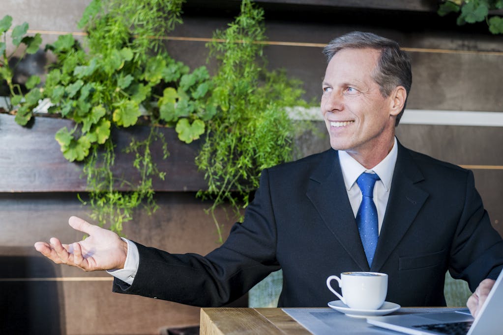 Smiling mature businessman making hand gesture with cup of coffee and laptop on desk