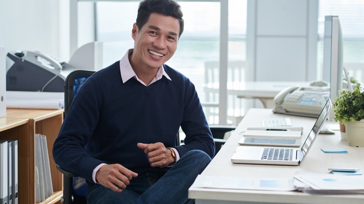 Smiling man sitting at desk in front of laptop in office and looking at camera