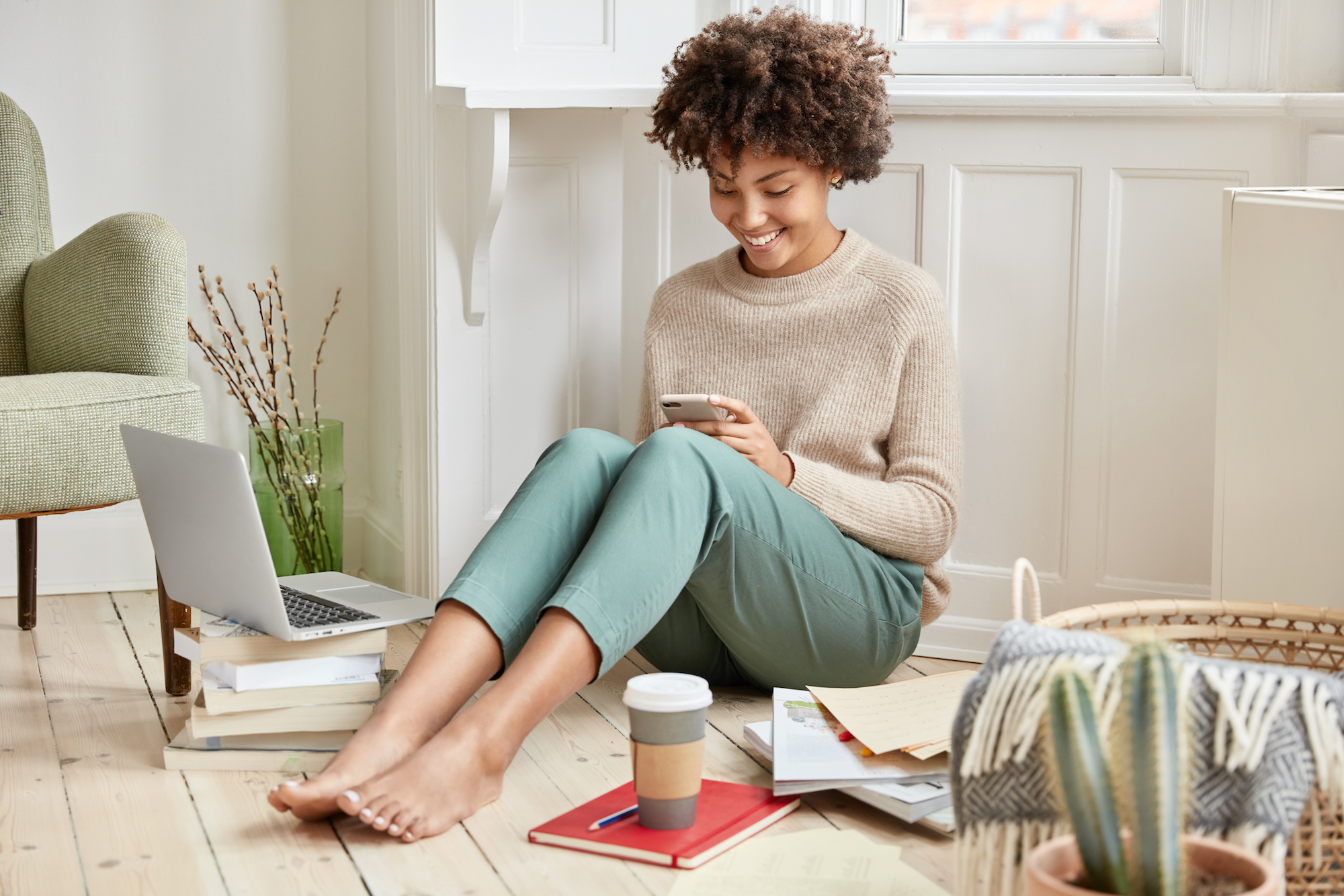 Positive young woman with afro haircut, dressed in fashionable clothes looking at her cell phone while smiling. <a href='https://www.freepik.com/photos/coffee'>Coffee photo created by wayhomestudio - www.freepik.com</a>