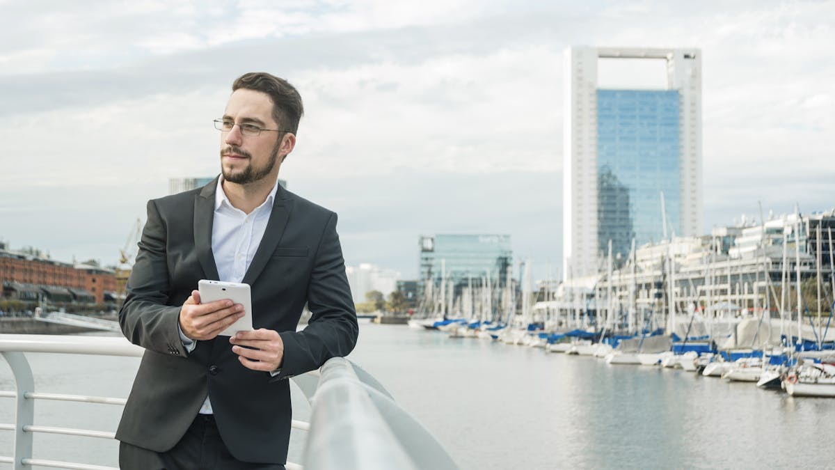 Retrato de un joven hombre de negocios de pie cerca del puerto sosteniendo un teléfono móvil