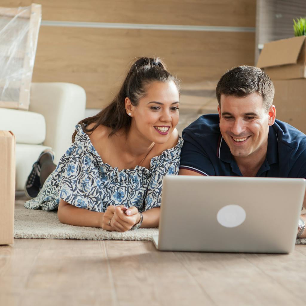 Husband and wife on their laptop in the living room of their home surrounded by moving boxes