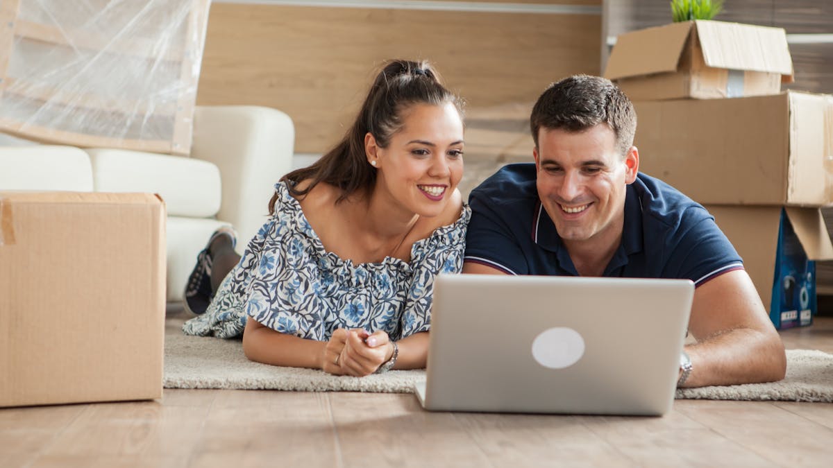 Husband and wife on their laptop in the living room of their home surrounded by moving boxes