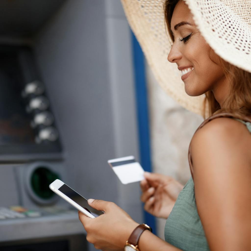 Smiling woman withdrawing money from cash machine while using smart phone