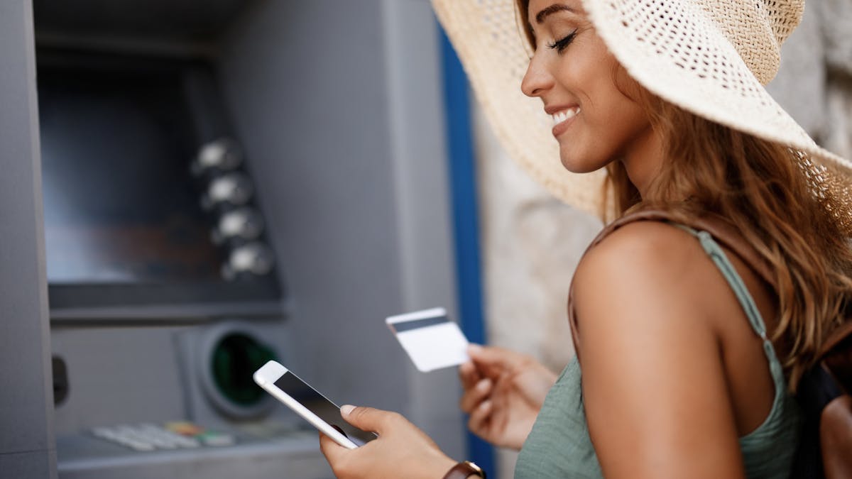 Smiling woman withdrawing money from cash machine while using smart phone