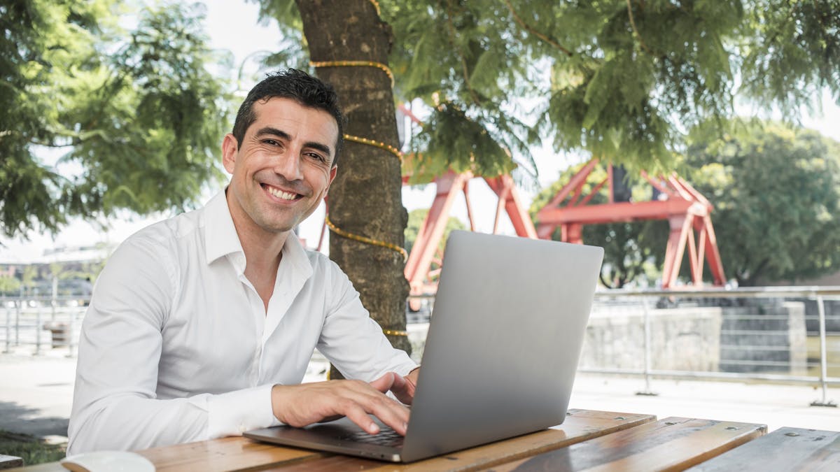 Joven emprendedor sonriendo al aire libre mientras trabaja en su computadora portátil