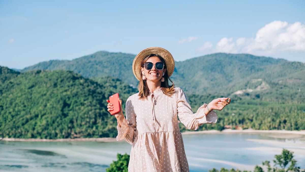 Young woman in summer cute dress, straw hat and sunglasses dancing with smartphone on hand