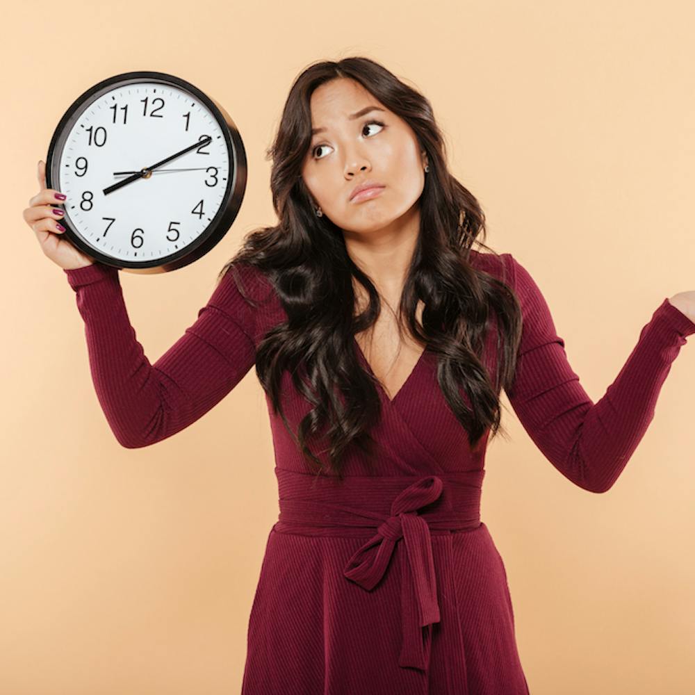 Puzzled brunette woman with curly long hair holding clock showing time after 8 gesturing like she is late but not worried, over peach background