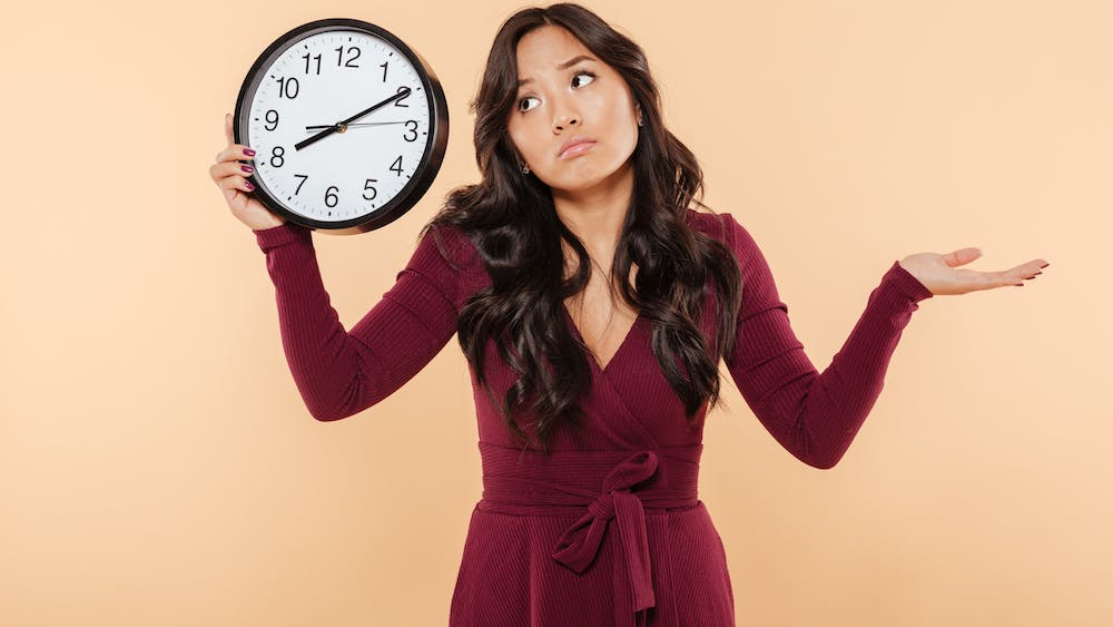 Puzzled brunette woman with curly long hair holding clock showing time after 8 gesturing like she is late but not worried, over peach background