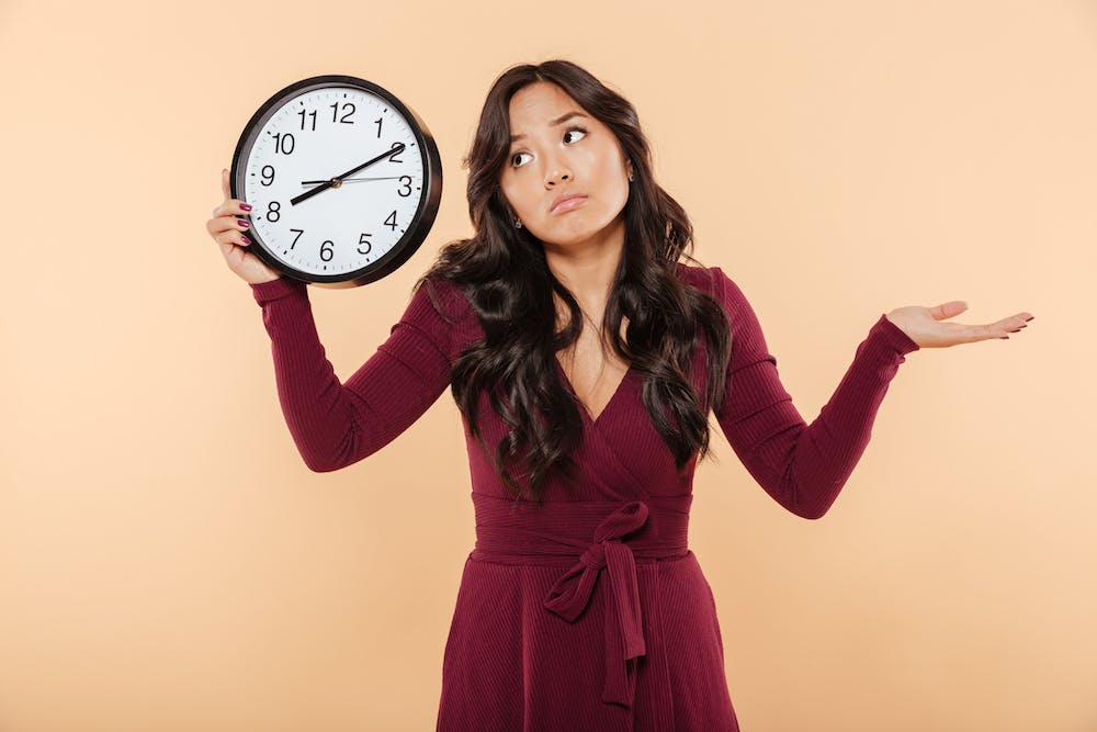 Puzzled brunette woman with curly long hair holding clock showing time after 8 gesturing like she is late but not worried, over peach background