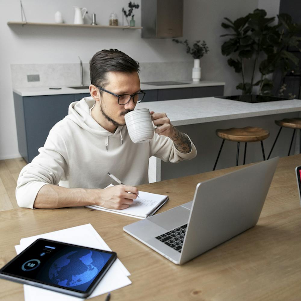 Photo of a man working from home at desk while having a coffee. Image by <a href="https://www.freepik.com/free-photo/man-working-from-home-desk-while-having-drink_21076515.htm#page=2&query=remote%20worker&position=19&from_view=search&track=ais">Freepik</a>