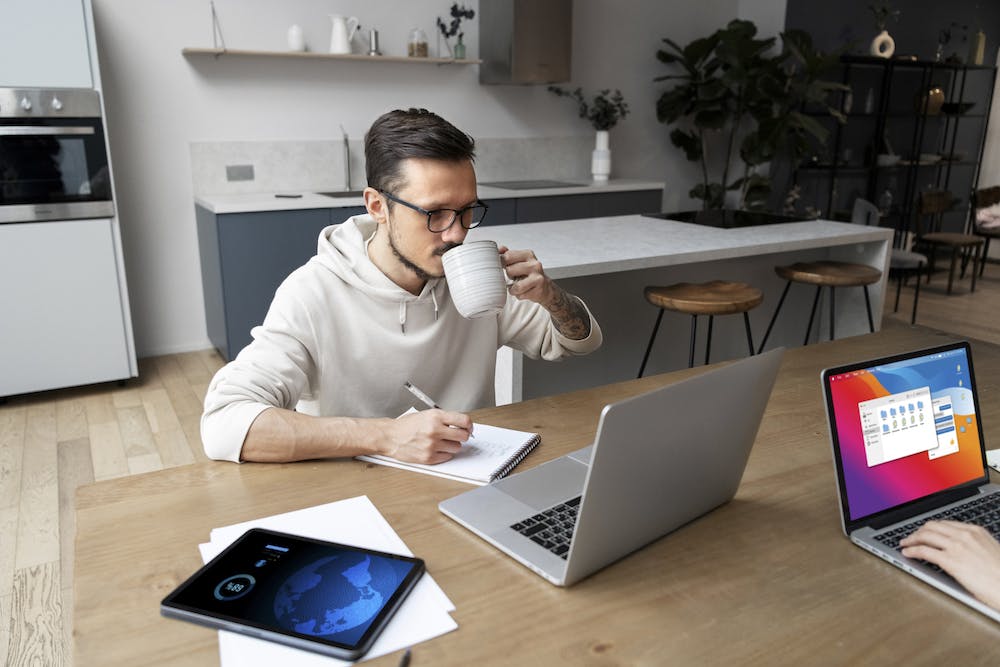 Photo of a man working from home at desk while having a coffee. Image by <a href="https://www.freepik.com/free-photo/man-working-from-home-desk-while-having-drink_21076515.htm#page=2&query=remote%20worker&position=19&from_view=search&track=ais">Freepik</a>