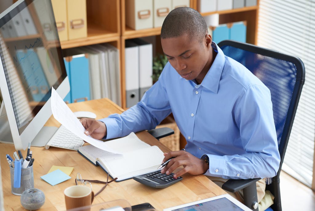 Man with blue collar shirt using a calculator with a sheet of paper in right hand