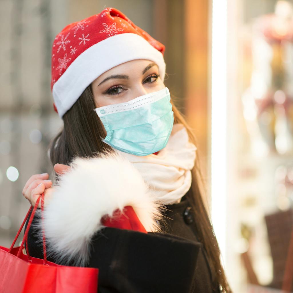 Woman wearing a red holiday hat and a mask with holiday lights on the background