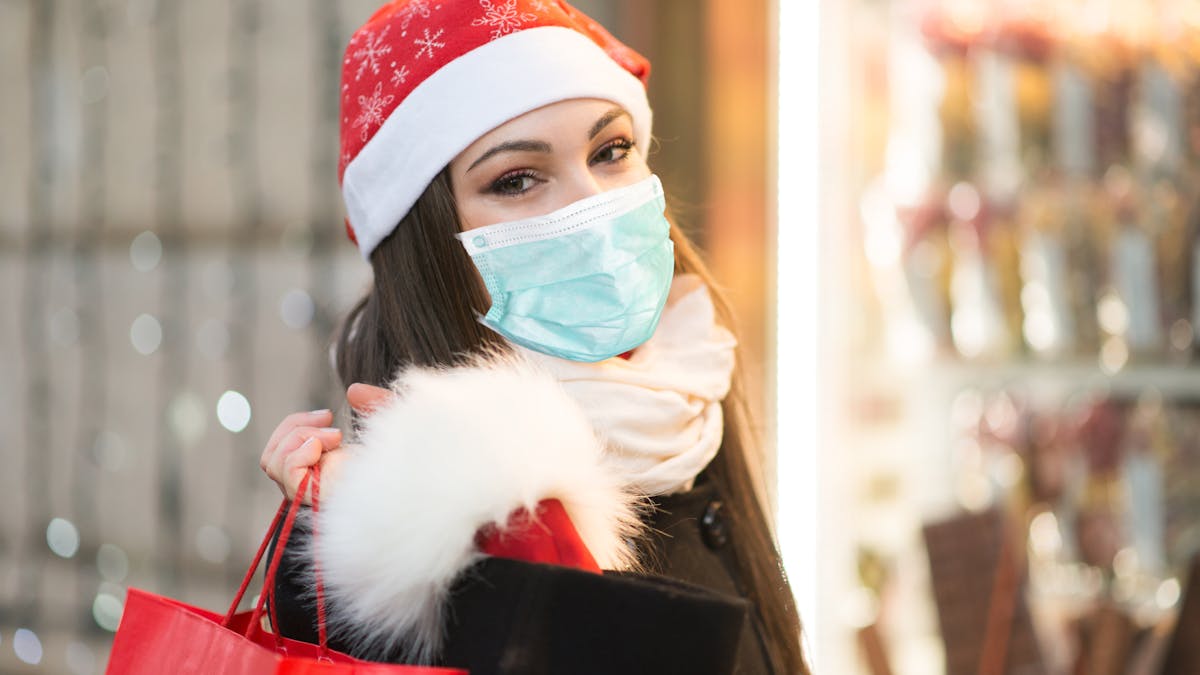 Woman wearing a red holiday hat and a mask with holiday lights on the background