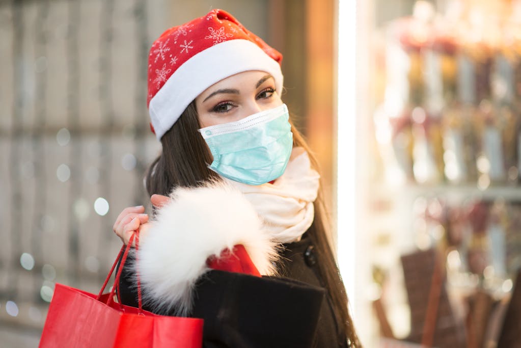 Woman wearing a red holiday hat and a mask with holiday lights on the background