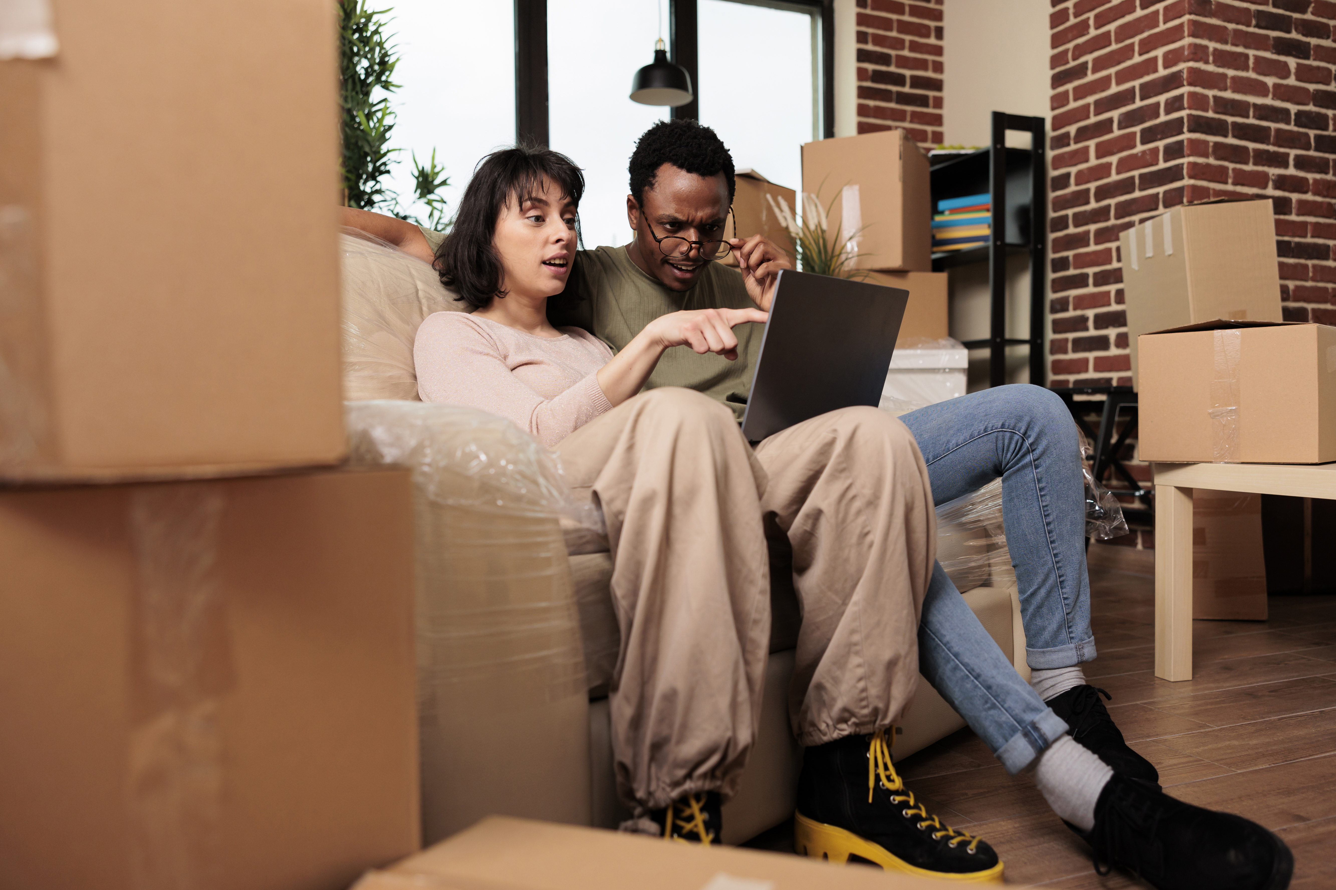 A couple sitting on top of moving boxes looking intently at a laptop.
