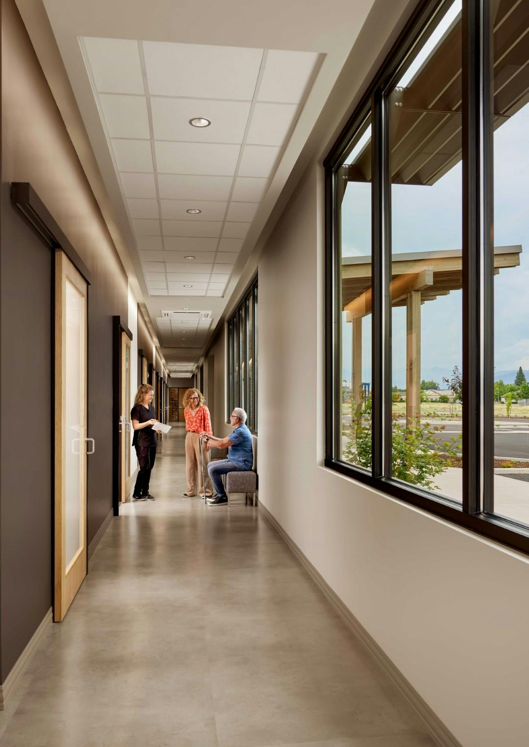 The outer corridor of the OOC ambulatory surgery center. A patient and their family member are talking to a nurse. The hallway is very well lit due to the natural lighting of the large windows.