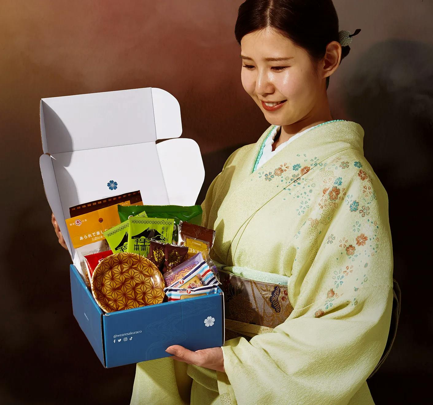 A woman in a kimono stands holding Sakuraco's May box